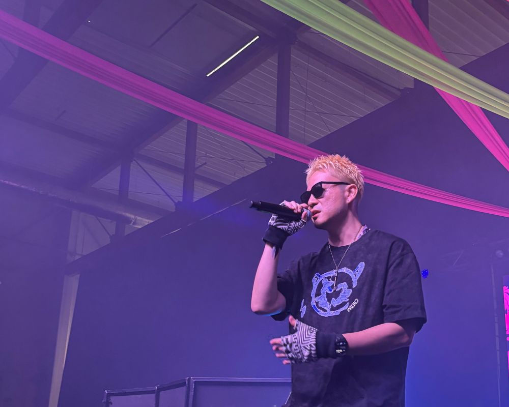 Na zdjęciu widzimy rapera Lotus Juice'a. Mężczyzna ma blond włosy. Ma na sobie okulary przeciwsłoneczne, luźny, czarny t-shirt, czarną bandanę na nadgarstku oraz biało-czarne rękawiczki. W prawej ręce trzyma mikrofon. W tle widzimy halę, w której odbywał się koncert. Przyozdobiono ją kolorowymi szarfami (różowe i zielone) oraz światłami (tutaj fioletowo-niebieskie).
