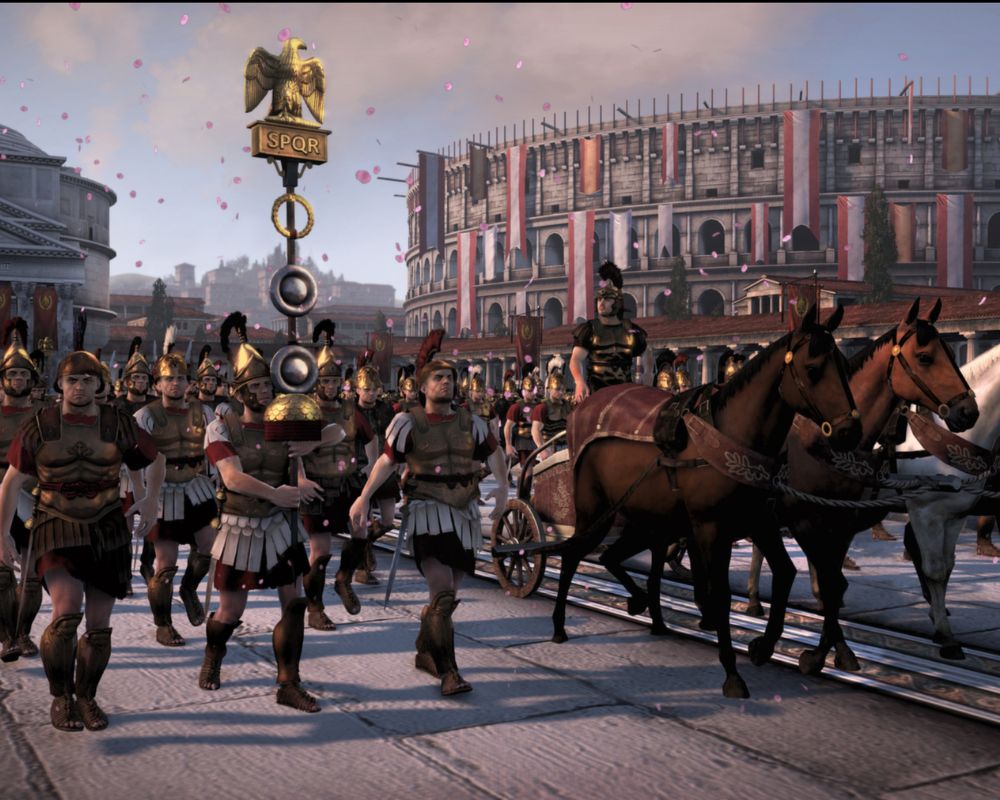 Zrzut ekranu z gry Total War: Rome II przedstawiający uroczysty triumf rzymski na placu miejskim. W centrum żołnierze legionu w lśniących, złotych zbrojach i hełmach z pióropuszami maszerują obok rydwanu zaprzężonego w dwa ciemne konie. Jeden z legionistów dumnie dzierży wysoki rzymski znak legionowy (Akwila) zwieńczony złotym orłem i tabliczką z napisem „SPQR”. W tle góruje monumentalny amfiteatr przypominający Koloseum, udekorowany czerwono-białymi sztandarami. Z nieba sypią się różowe płatki kwiatów, nadając scenie podniosły, zwycięski charakter. Słońce rzuca długie cienie na kamienny bruk.