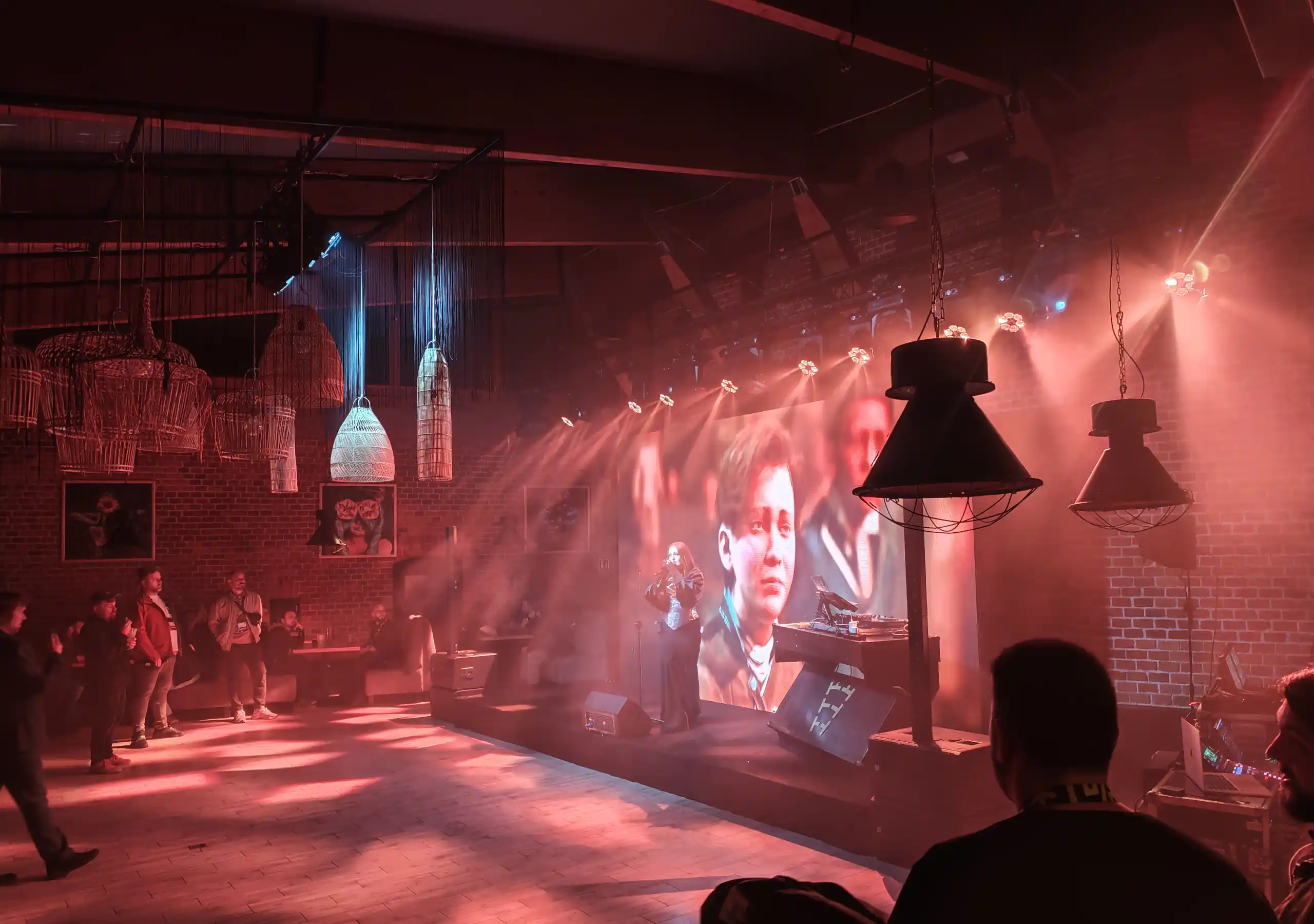 Zdjęcie przedstawia występ muzyczny na żywo, odbywający się w klimatycznym, industrialnym klubie lub sali eventowej o ceglanych ścianach, zdominowanej przez ciepłe, czerwone oświetlenie i efekty mgły. Na scenie w tle widoczna jest wokalistka występująca przed wielkim ekranem LED, na którym wyświetlane są bliskie ujęcia twarzy postaci (wyglądających jak kadry z gry wideo lub filmu), a obok niej, za konsolą didżejską, znajduje się stanowisko muzyczne. Estrada jest oświetlona silnymi, ukośnymi snopami światła z reflektorów zamontowanych pod sufitem, które tworzą dynamiczne smugi w powietrzu i rzucają czerwone blaski na drewnianą podłogę przed sceną. Po lewej stronie kadru, w głębi sali, stoi niewielka grupa rozproszonych uczestników obserwujących występ, nad którymi wiszą dekoracyjne, plecione klosze lamp, natomiast na pierwszym planie po prawej stronie widoczne są sylwetki osób z publiczności (w tym mężczyzna tyłem) oraz fragmenty sprzętu realizatorskiego.