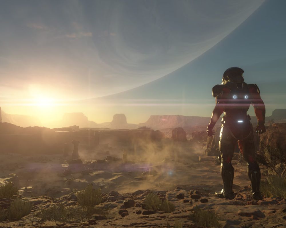 Zrzut ekranu z gry Mass Effect: Andromeda, widzimy na nim krajobraz jednej z planet. Jest pustynny, nad piaszczysto-skalną powierzchnią unosi się kurz, w tle widać wschodzące lub zachodzące słońce, a także zarys jakiejś innej planety lub gwiazdy. Na pierwszym planie, tyłem do widza, nieco z boku stoi postać w bojowym skafandrze kosmicznym. Na plecach znajdują się trzy świecące na niebiesko lampki