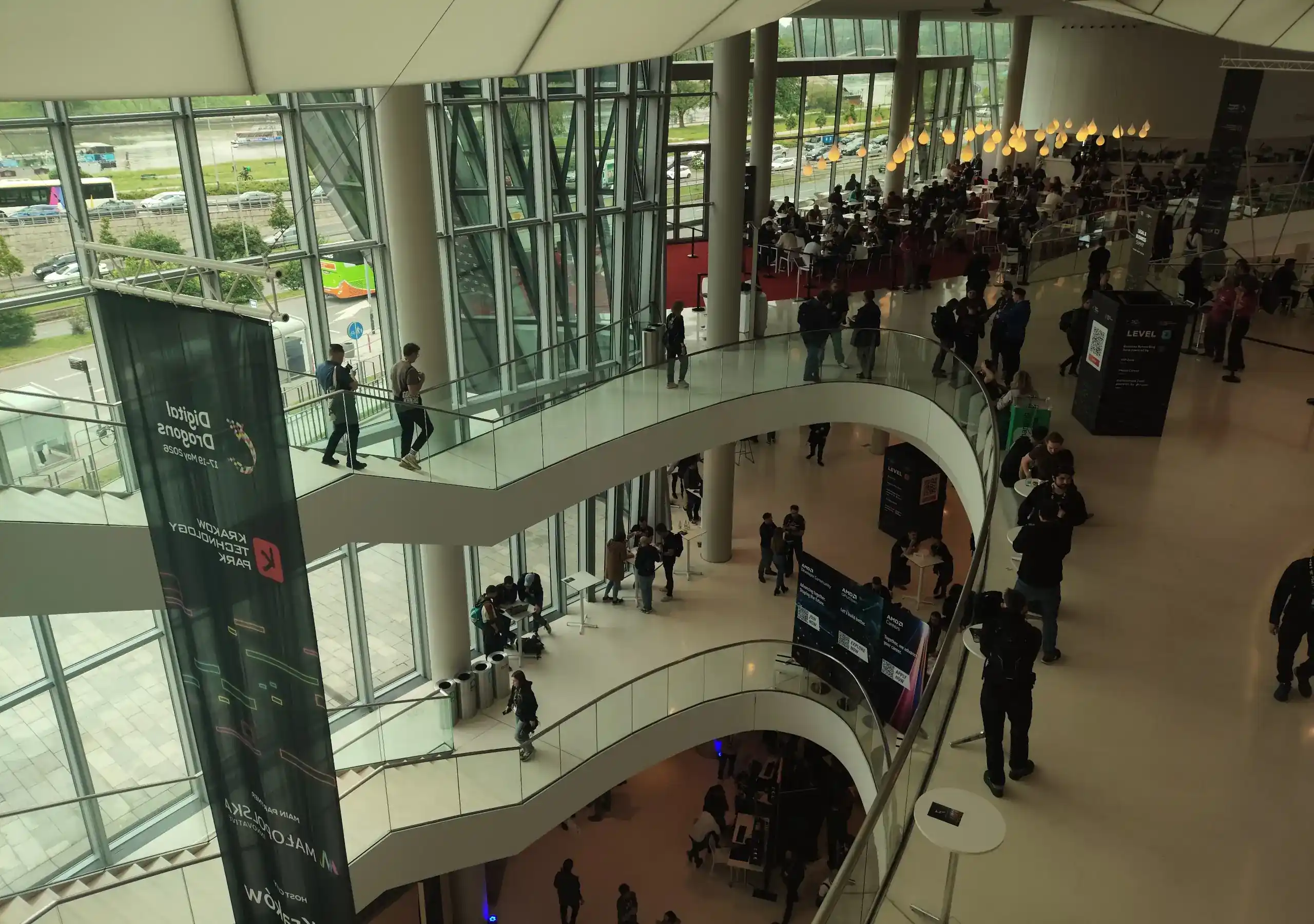Zdjęcie przedstawia z góry panoramiczny widok na nowoczesne, wielopoziomowe wnętrze przeszklonego budynku (Centrum Kongresowe ICE Kraków) podczas wydarzenia branżowego, na co wskazuje wielki, pionowy baner z logo „Digital Dragons” i „Krakow Technology Park” zwisający po lewej stronie. Architektura obiektu charakteryzuje się falistymi, białymi schodami i korytarzami z szklanymi balustradami, które łączą poszczególne piętra, a ogromne, panoramiczne okna odsłaniają widok na zewnętrzną ulicę, rzekę oraz przejeżdżające samochody i autobusy. Na różnych poziomach widać rozproszone grupy ludzi – uczestników konferencji spacerujących po korytarzach, rozmawiających przy stolikach koktajlowych lub gromadzących się w strefach informacyjnych i gastronomicznych, gdzie w głębi po prawej stronie rozwinięto czerwony dywan. Wnętrze jest jasne, oświetlone naturalnym światłem wpadającym przez fasadę oraz ozdobnymi, wiszącymi lampami w kształcie kropel, tworząc przestronną i dynamiczną atmosferę masowego wydarzenia technologicznego.