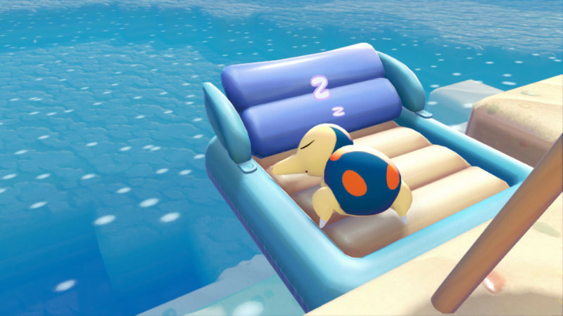 Pokemon Cyndaquill spokojnie śpi na dmuchanym materacu. Materac unosi się na wodach oceanu, w zasięgu tła widać także kawałek plaży.
