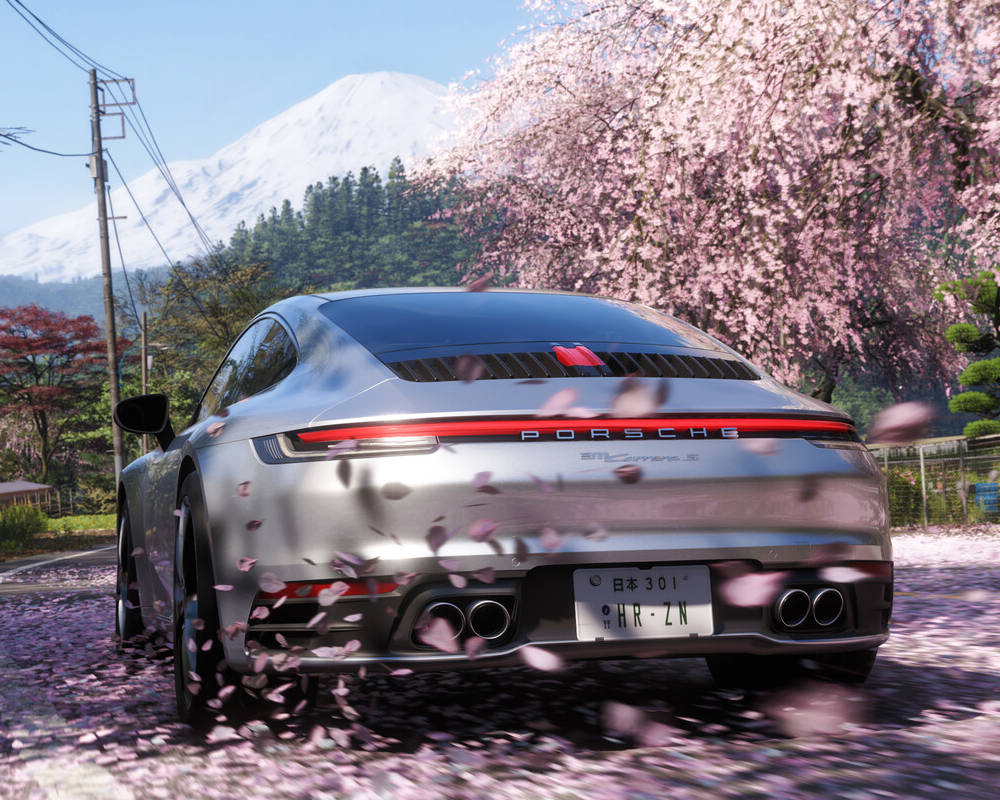 Ten zrzut ekranu pochodzi bezpośrednio z gry i przedstawia tylną część sportowego samochodu marki Porsche (model 911 Carrera S) w kolorze srebrnym. Pojazd jedzie po asfaltowej drodze, a spod jego kół dynamicznie unoszą się płatki różowej wiśni. Sceneria ma wyraźnie japoński klimat. Po prawej stronie drogi rośnie duże, pięknie kwitnące drzewo sakura (wiśnia), z którego opadają wspomniane płatki. W tle, po lewej stronie, wznosi się majestatyczna, pokryta śniegiem góra, przypominająca górę Fudżi. Całość uzupełnia japońska tablica rejestracyjna na aucie oraz tradycyjne słupy telegraficzne, co jednoznacznie wskazuje na miejsce akcji nowej odsłony gry. Kadr jest niezwykle fotorealistyczny, z pięknym oświetleniem i rozmyciem ruchu.