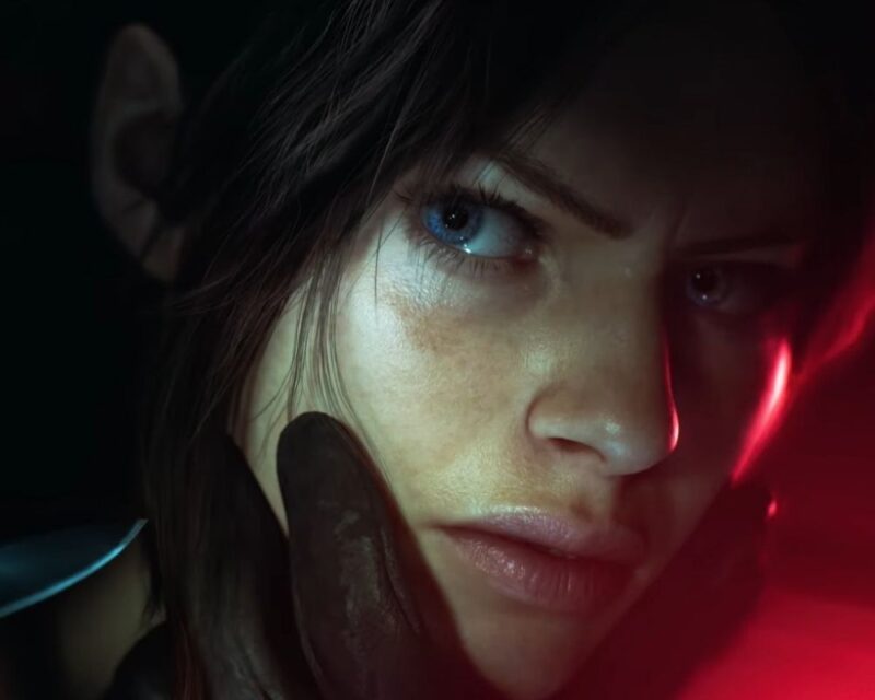 Grafika przedstawia zbliżenie na twarz Claire Redfield z nadchodzącego remake'u Resident Evil Code: Veronica. Bohaterka ma ciemne włosy i niebieskie oczy, a jej twarz oświetlają kontrastujące czerwone i chłodne światła, podkreślające poważny, skupiony wyraz twarzy.
