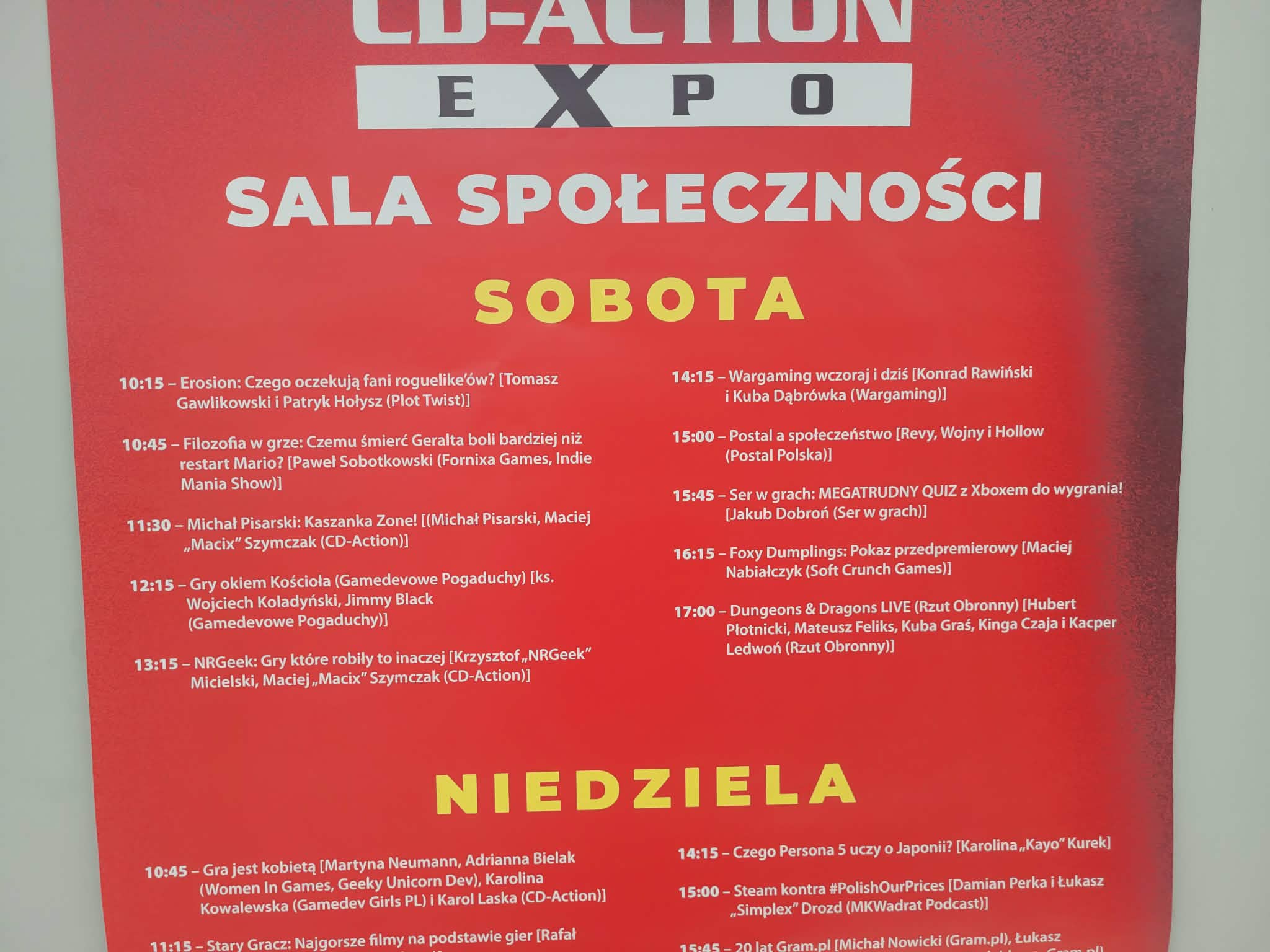 Czerwony plakat z wydarzenia CD-Action Expo przedsstawiający wydarzenia odbywające się w sali społeczności. Widoczny ejst harmonogram na dwa dni wydarzenia i nazwy odbywających się wykładów