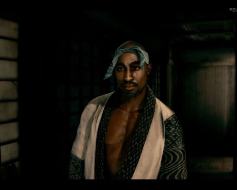 Tupac Shakur przedstawiony w postaci z gry wideo stoi w słabo oświetlonym, tradycyjnym japońskim wnętrzu. Ma ogoloną głowę, krótki zarost oraz jasną chustę zawiązaną wokół czoła. Ubrany jest w luźne kimono w ciemne wzory z jasnymi panelami po bokach, odsłaniające część klatki piersiowej. Mężczyzna patrzy lekko w bok z poważnym, spokojnym wyrazem twarzy. Tło stanowi ciemny korytarz z drewnianymi ścianami i przesuwanymi panelami, oświetlony ciepłym, przytłumionym światłem. Kadr pochodzi z gry wideo i utrzymany jest w realistycznej stylistyce.