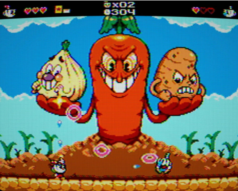 Obrazek przedstawia zrzut ekranu z retro gry platformowej stylizowanej na produkcję z ery 8-bitowej, nawiązującej bezpośrednio do walki z bossami „The Root Pack” z gry Cuphead. Całość wyświetlana jest z efektem przypominającym stary monitor CRT z widocznymi liniami skanowania. W centrum kadru dominuje wielka, antropomorficzna, pomarańczowa marchewka o złowrogim, szerokim uśmiechu z odsłoniętymi zębami i groźnym spojrzeniu, która wyrasta prosto z ziemi. Postać ta trzyma w swoich dłoniach-liściach dwóch mniejszych bossów: po lewej stronie płaczącą, białą cebulę o smutnym wyrazie twarzy, a po prawej zagniewanego, brązowego ziemniaka o zaciśniętych zębach. Na dole ekranu, na brązowym podłożu usianym drobnymi roślinami, widoczne są dwie małe postacie grywalnych bohaterów – Cuphead po lewej i Mugman po prawej stronie – którzy unikają unoszących się w powietrzu różowych i niebieskich pocisków lub pierścieni. Tłem dla całej sceny jest jasnoniebieskie niebo z prostymi, pikselowymi chmurami, a na samej górze ekranu umieszczono klasyczny interfejs gracza (HUD) wyświetlający ikony postaci, serduszka oznaczające punkty życia, licznik żyć („x02”) oraz punkty („304”).