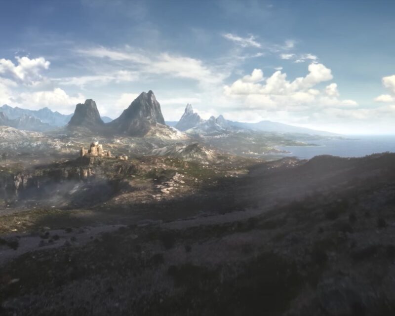 Szeroki kadr z lotu ptaka przedstawiający surowy, górzysty krajobraz fantasy ze zwiastuna The Elder Scrolls VI. Na pierwszym planie widać zacienione, skaliste wzgórza spowite lekką mgłą. W środkowej części, na skalnym wzniesieniu po lewej stronie, wznoszą się ruiny kamiennej fortecy lub zamku. W tle dominują wysokie, ostre szczyty górskie, a po prawej stronie linia brzegowa przechodzi w otwarte, niebieskie morze. Całość oświetla jasne słońce przedzierające się przez białe chmury na błękitnym niebie.
