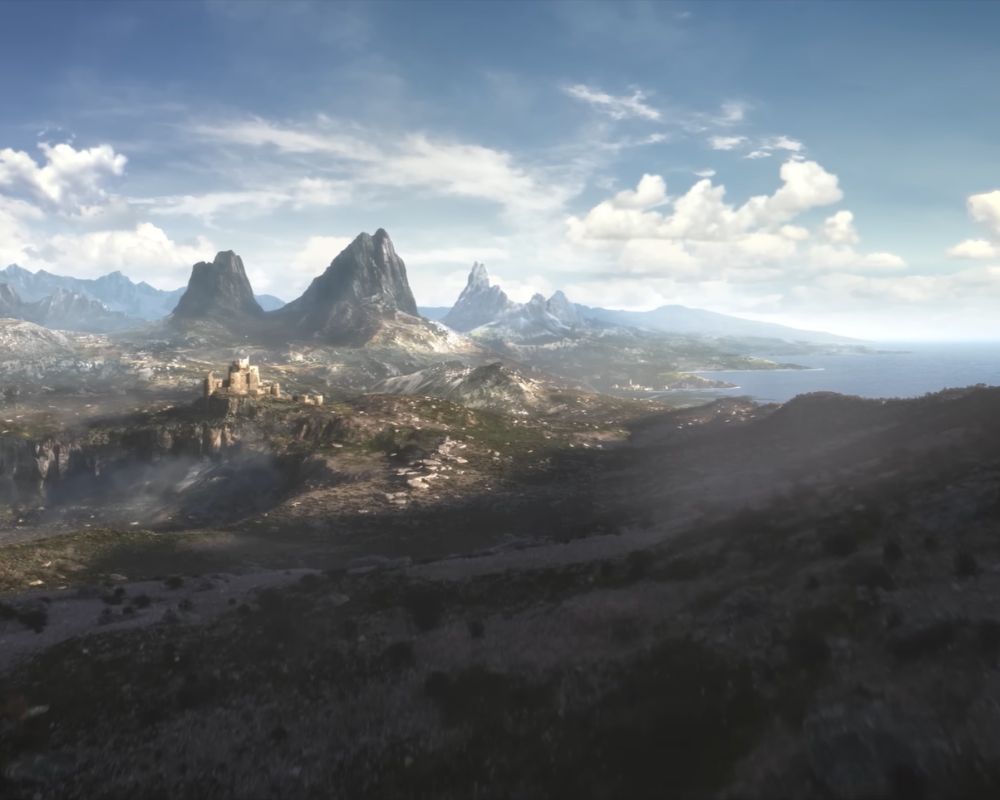 Szeroki kadr z lotu ptaka przedstawiający surowy, górzysty krajobraz fantasy ze zwiastuna The Elder Scrolls VI. Na pierwszym planie widać zacienione, skaliste wzgórza spowite lekką mgłą. W środkowej części, na skalnym wzniesieniu po lewej stronie, wznoszą się ruiny kamiennej fortecy lub zamku. W tle dominują wysokie, ostre szczyty górskie, a po prawej stronie linia brzegowa przechodzi w otwarte, niebieskie morze. Całość oświetla jasne słońce przedzierające się przez białe chmury na błękitnym niebie.
