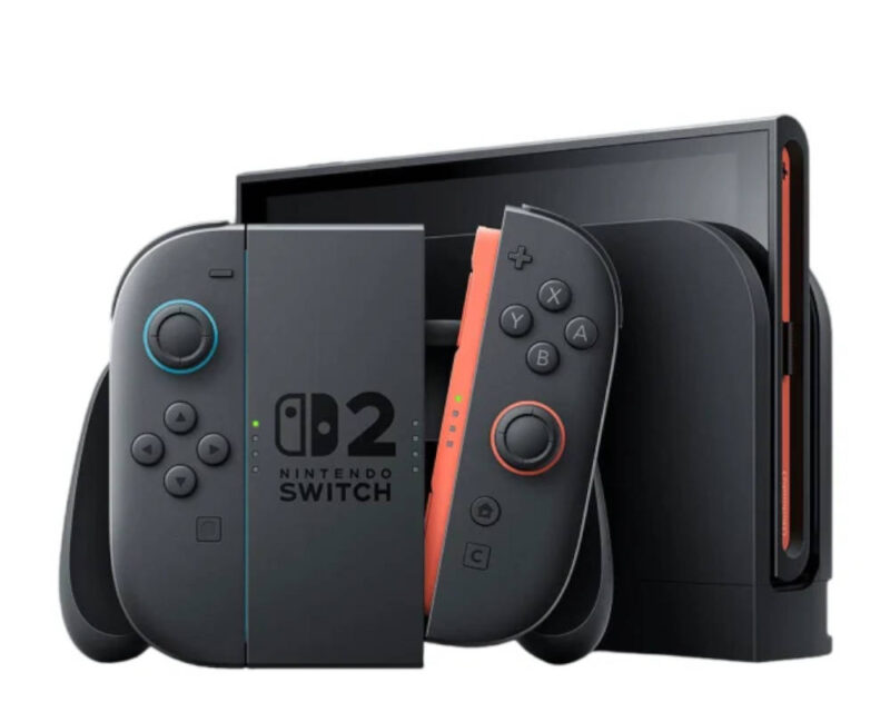 Obrazek przedstawia czarną konsolę do gier stylizowaną na hipotetycznego następcę Nintendo Switch, umieszczoną na białym tle. Na pierwszym planie po lewej stronie widoczny jest kontroler złożony z dwóch Joy-Conów wpiętych w uchwyt (grip), na którego środku nadrukowano logo Nintendo Switch z dużą cyfrą „2”. Kontrolery są ciemnoszare – lewy posiada analog z jasnoniebieskim akcentem i cztery przyciski kierunkowe, natomiast prawy ma analog z pomarańczowo-czerwonym akcentem, tradycyjne przyciski A/B/X/Y oraz widoczną pomarańczową krawędź boczną z zielonymi diodami LED. W tle, po prawej stronie, znajduje się sama konsola (ekran) włożona pionowo do czarnej stacji dokującej, z której boku wystaje czerwono-pomarańczowy element ozdobny lub gniazdo.